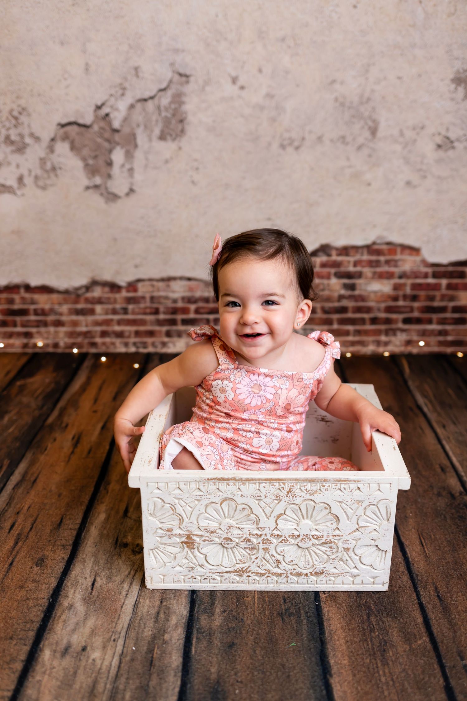 A pink dress-clad baby sits in a decorative white box against a rustic wooden floor and brick wall background.