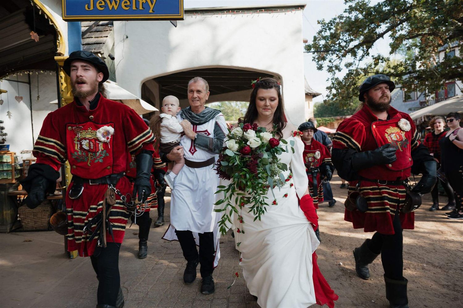 A wedding party in Renaissance attire proceeds through a medieval-themed marketplace.