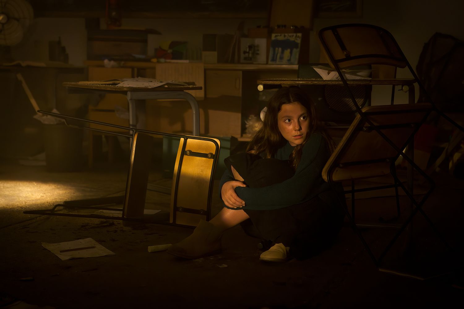 film still from a dramatic movie featuring a young girl hiding under a school desk with dramatic lighting