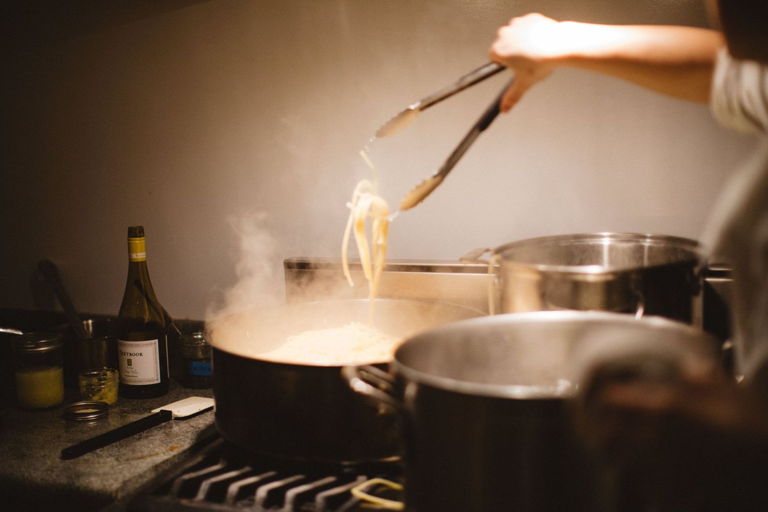 Steam rises as pasta is lifted from a large pot in a dimly lit kitchen.