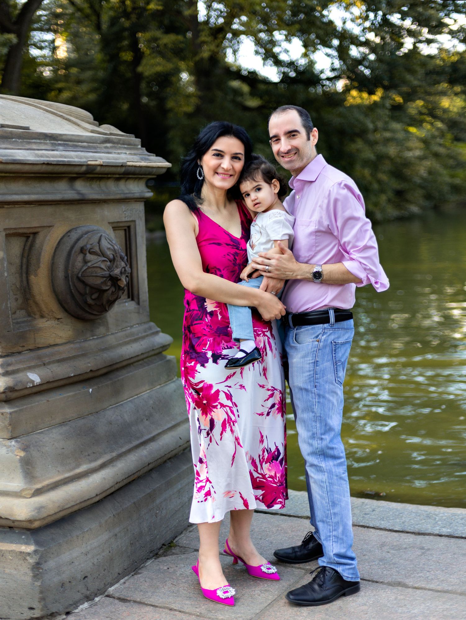 Family poses together by ornate stone railing near water in park setting.