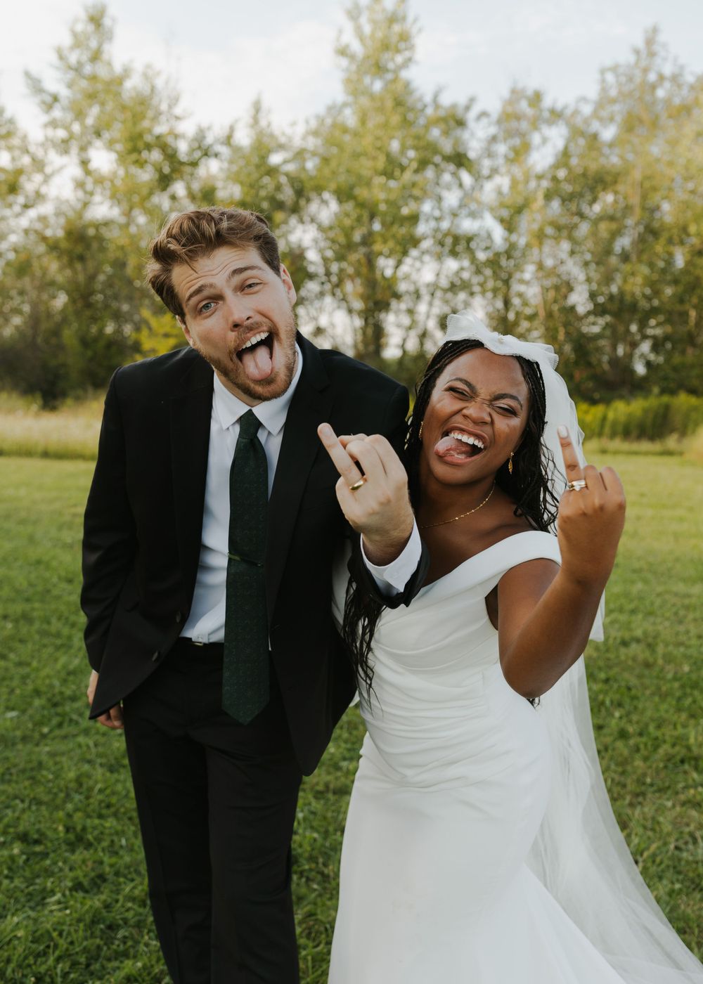 Joyful newlyweds make silly faces while posing in outdoor wedding photo.