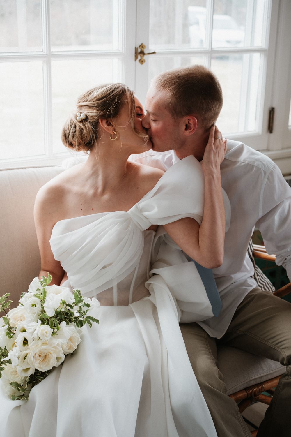 Romantic wedding moment with white dress and bouquet by bright windows.