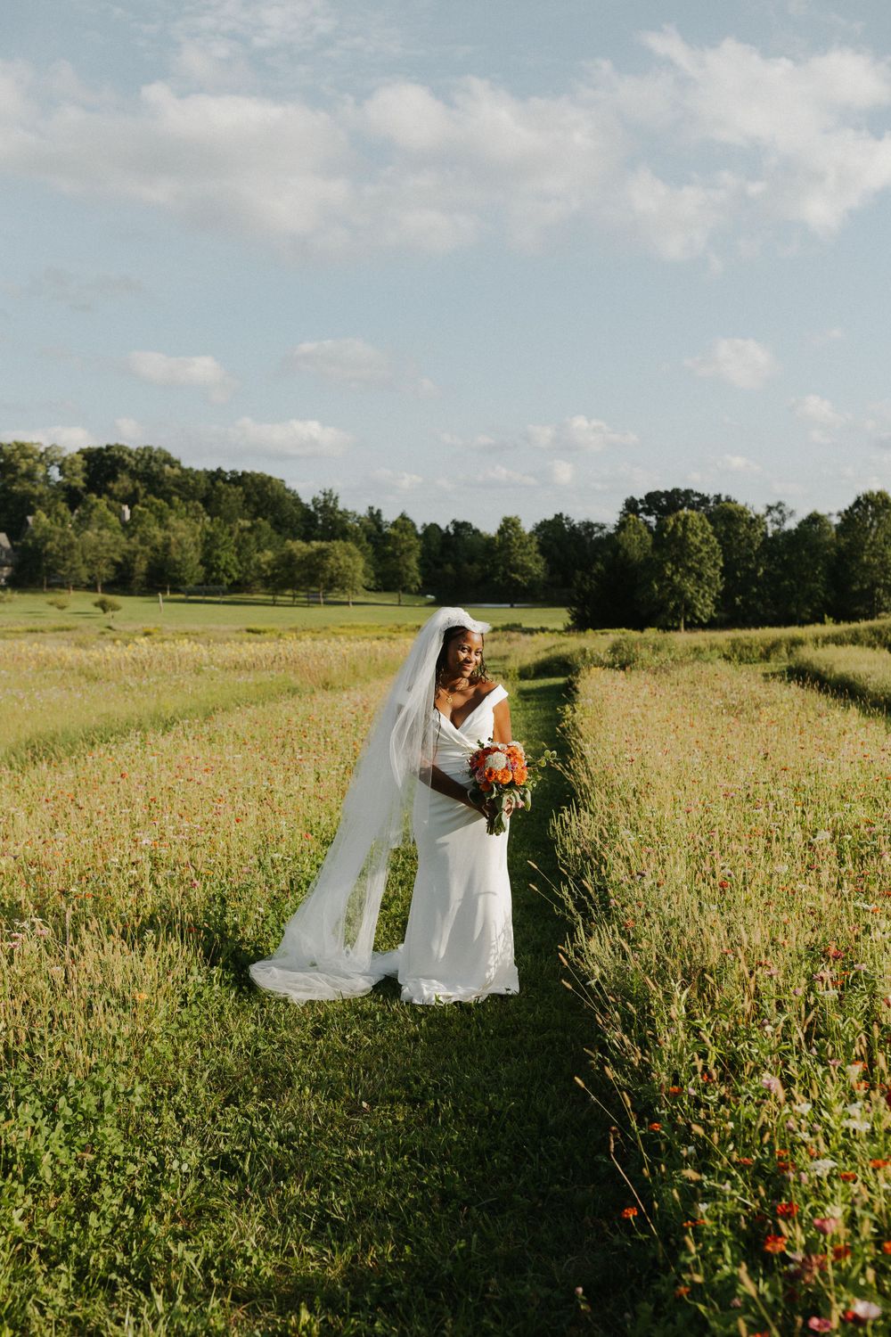 Bride stands in flowing white dress holding bouquet in sunlit wildflower meadow.