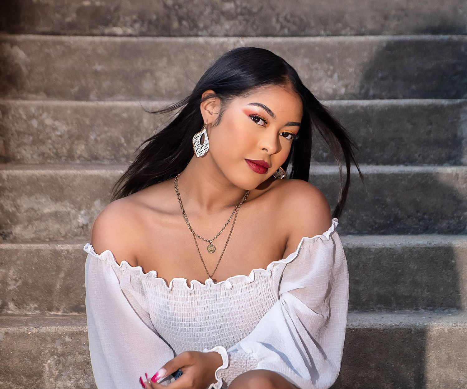 Portrait of woman in white off-shoulder top with jewelry against stone steps.