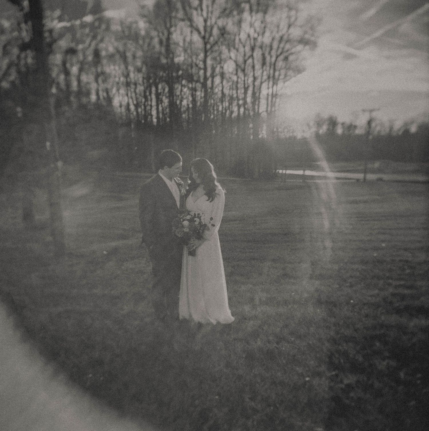 Bride and groom embracing at sunset in field, romantic black and white wedding portrait with lens flare.