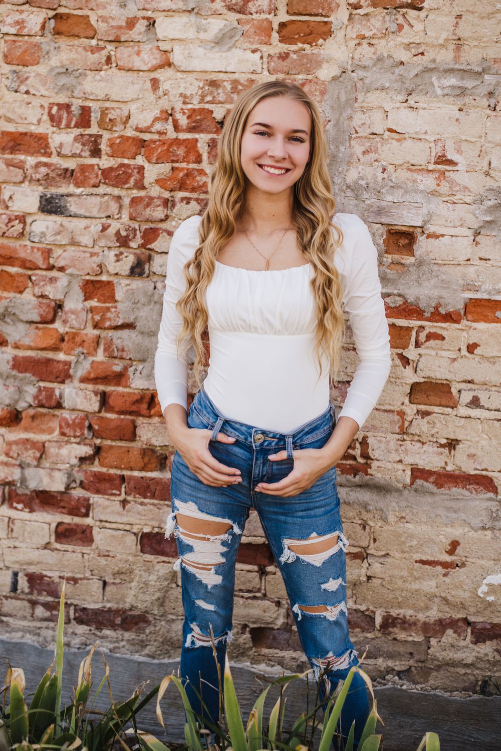 Casual fashion portrait with ripped jeans and white top against rustic brick background.