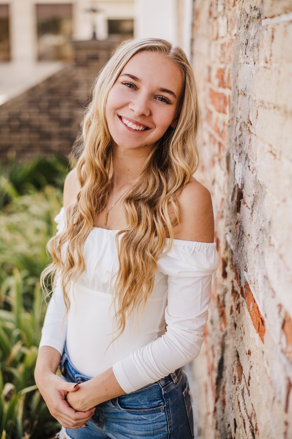 Portrait against weathered brick wall wearing white off-shoulder top and distressed denim in natural lighting.
