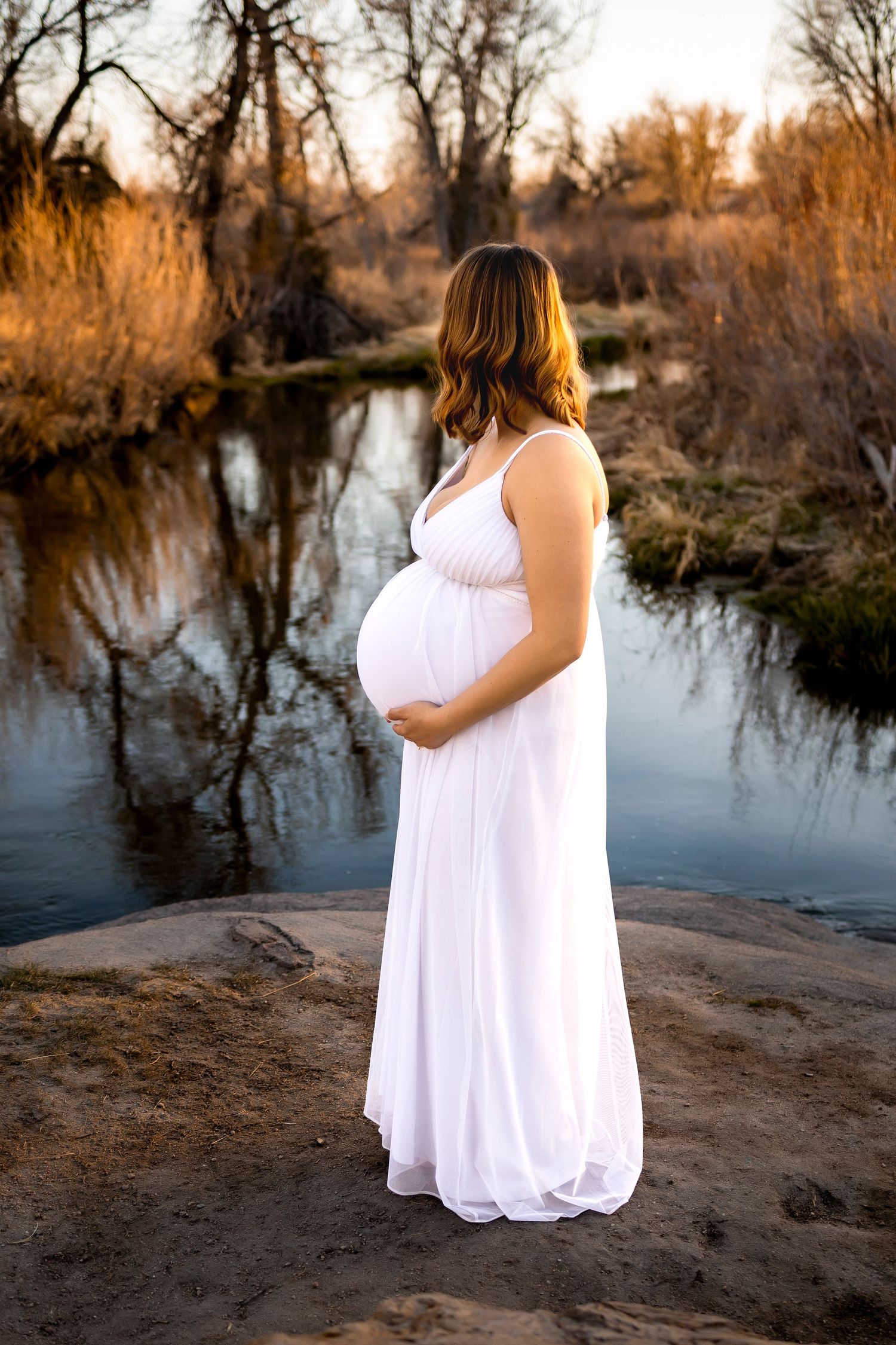 Pregnant woman in long white dress, she is looking away from camera towards a creek of water, long grass either side