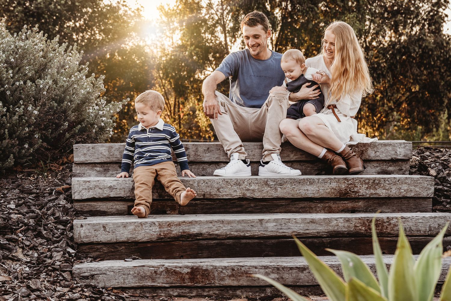 A family shares a relaxed moment together sitting on a wooden bench in a garden setting.