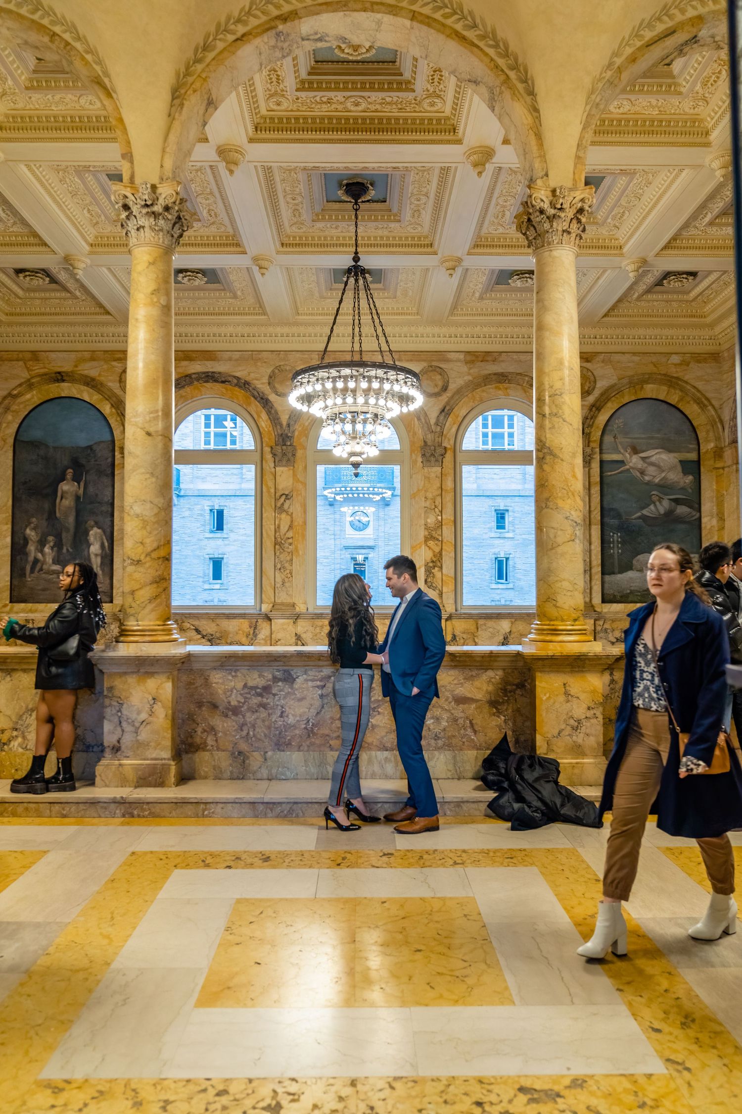 Ornate museum hall with marble columns, chandeliers and people walking through the grand space.