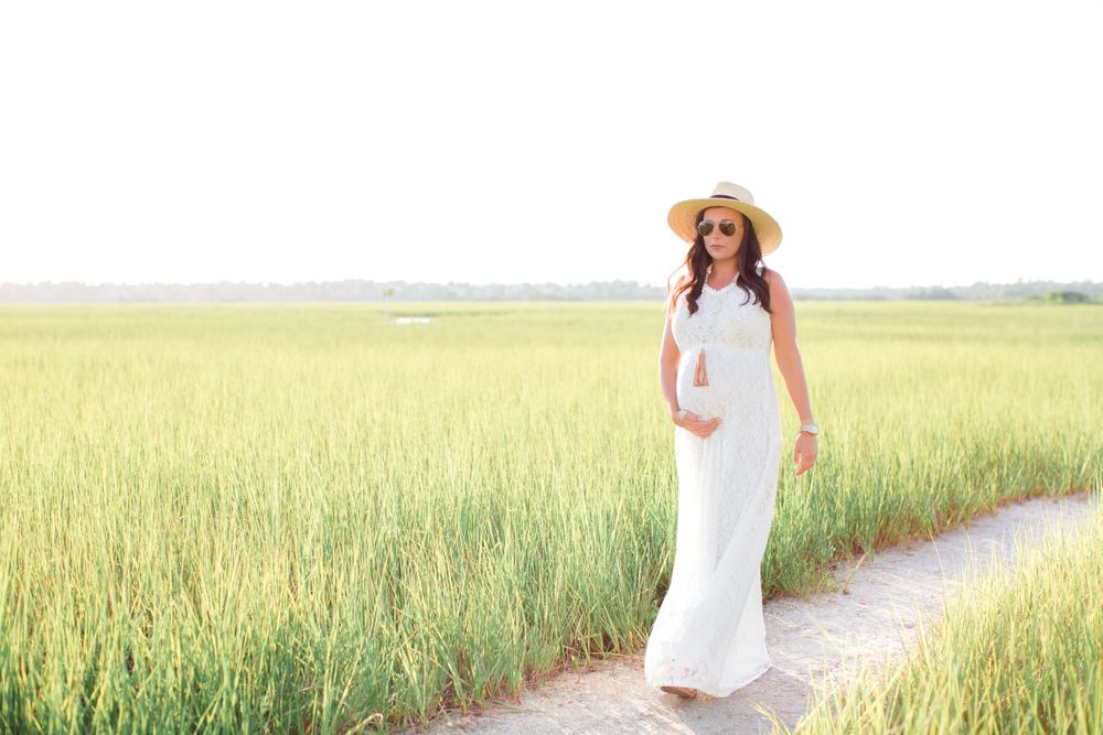 Maternity portrait of woman in white dress walking through golden grass field.