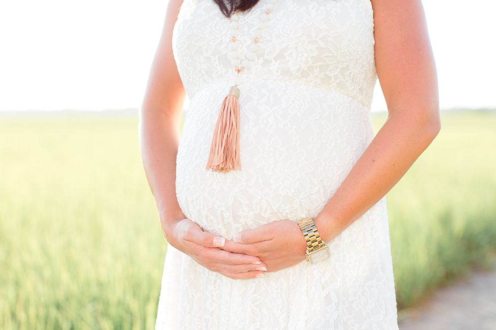 Detail shot of pregnant belly with tassel necklace against white lace maternity dress.