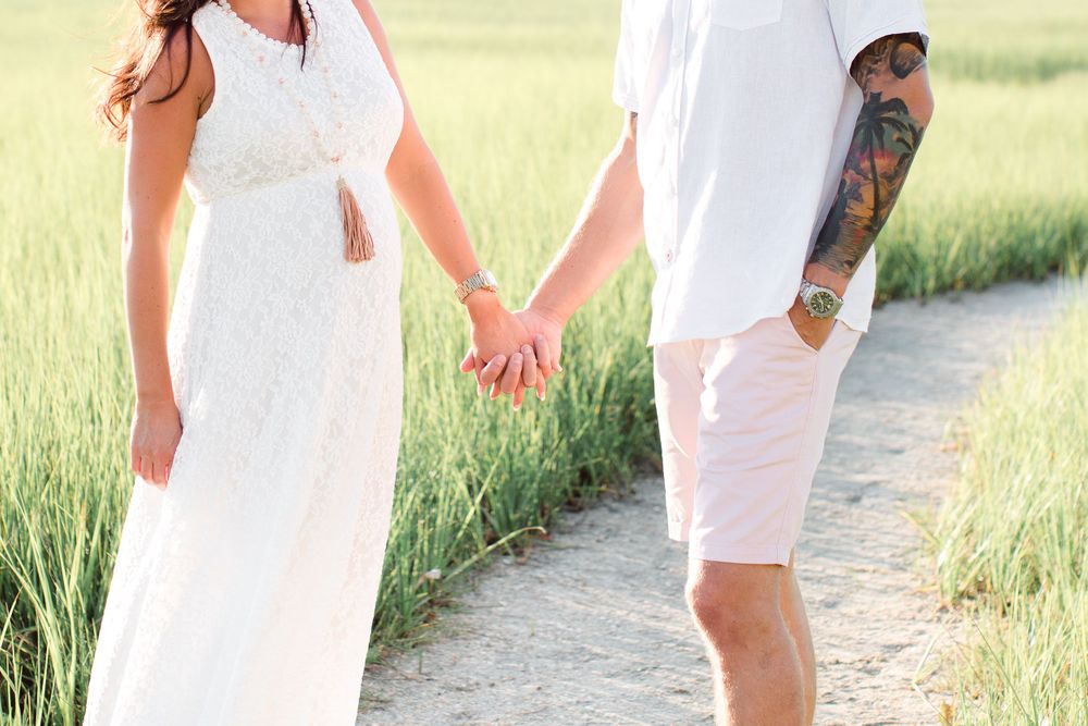 Couple holding hands on coastal path with marsh grass in background.