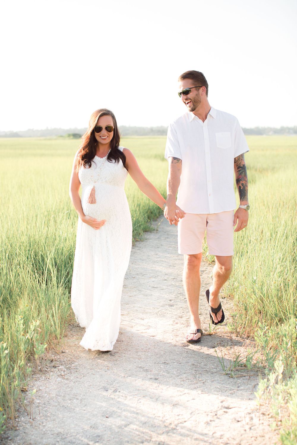Couple in white outfits holding hands while walking along a path through tall grass on a sunny day.
