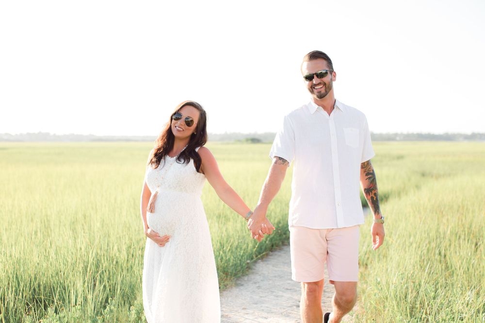 Couple dressed in white walking hand in hand through a grassy field on a bright summer day.