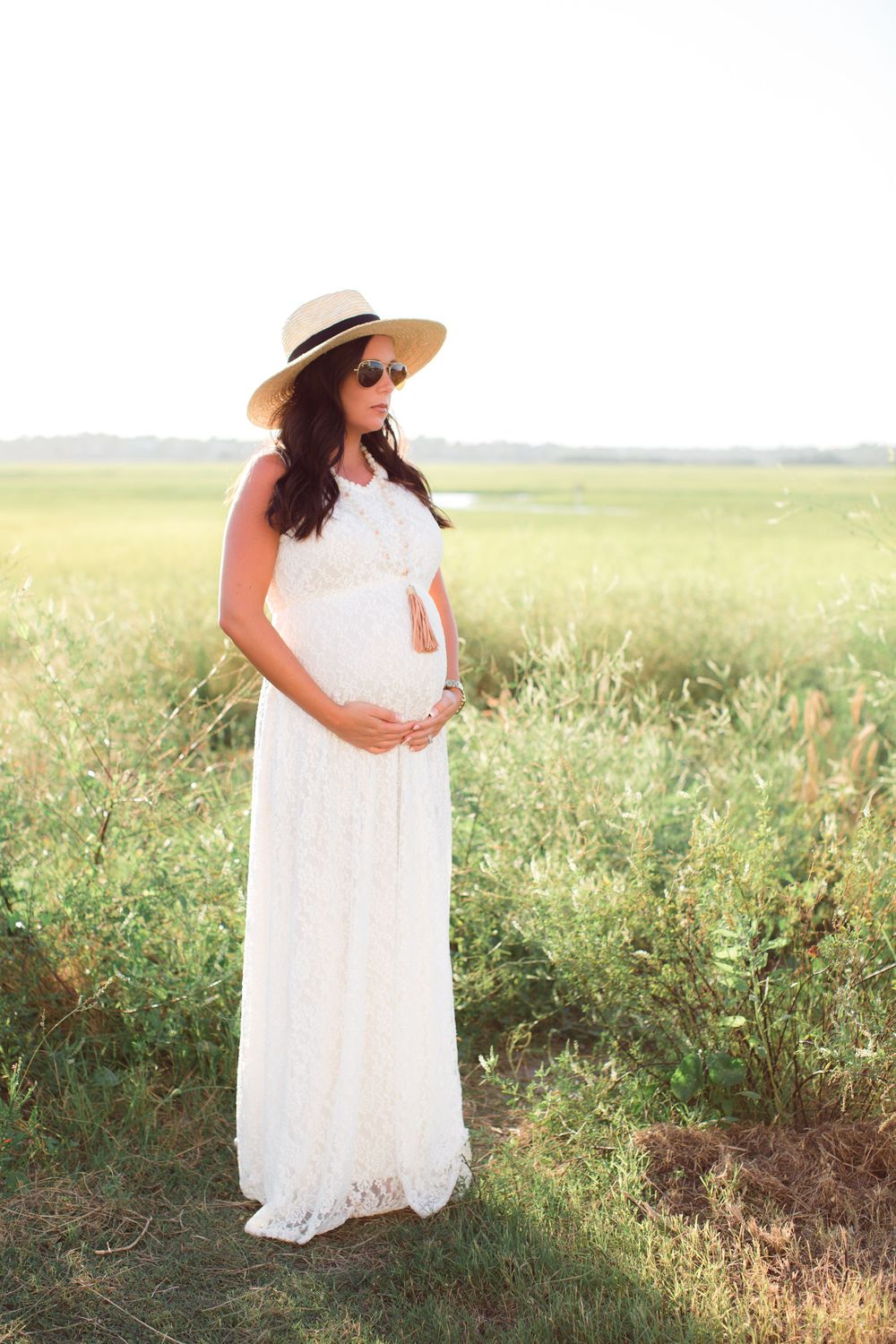 Woman in flowing white dress and sun hat poses in grassy field during sunset.
