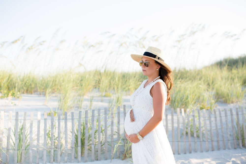 Woman in white dress and sun hat standing on beach with dunes and sea grass in background.