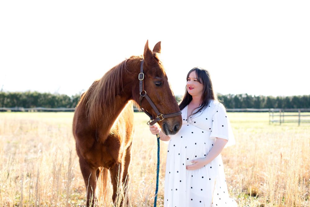 Woman in white polka dot dress poses with brown horse in golden field at sunset.