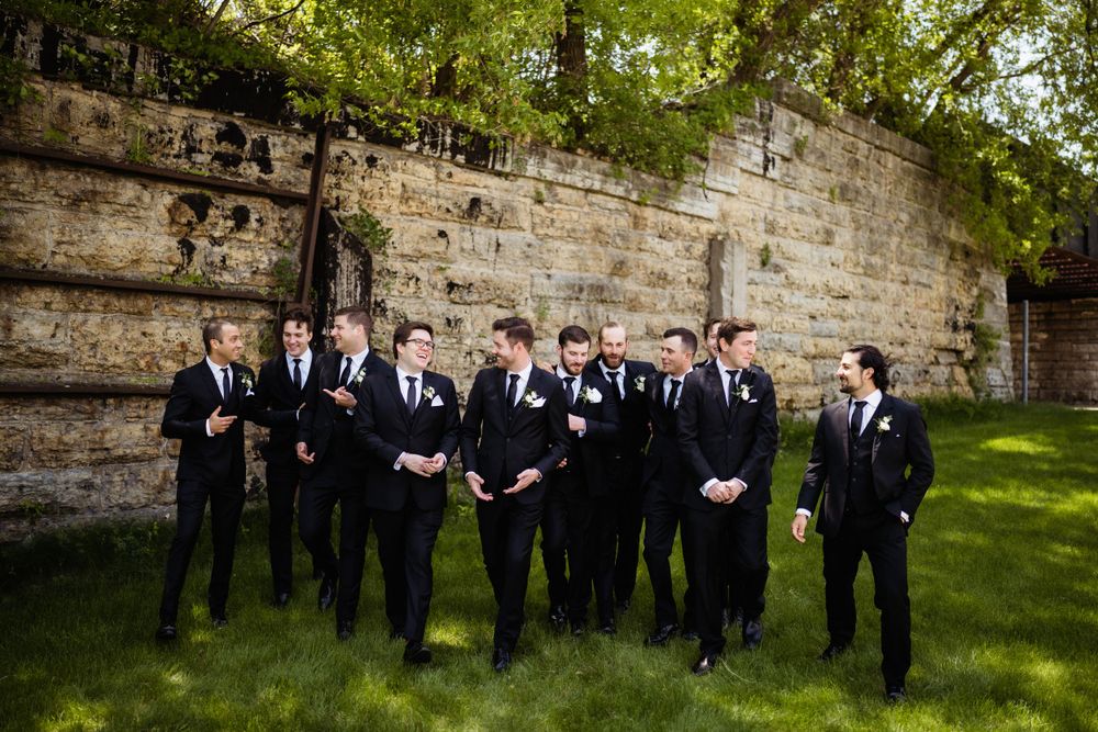 Groomsmen in black suits and ties pose formally against a stone wall outdoors.