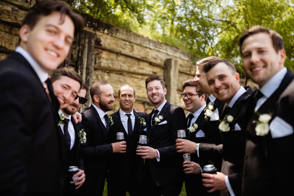 Groomsmen in formal attire share drinks and laughter during outdoor wedding photos.