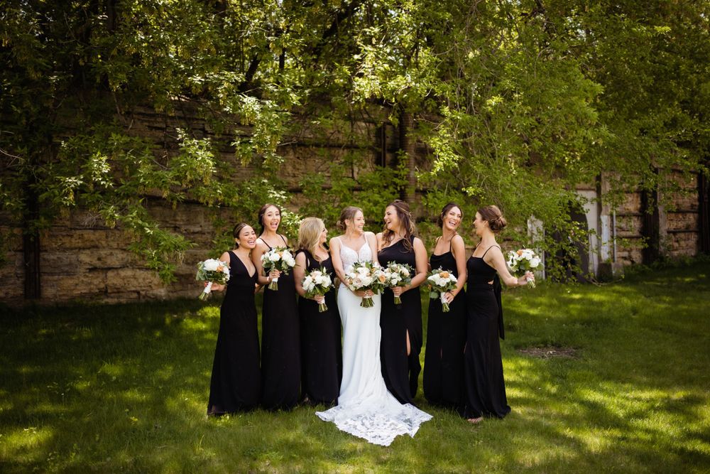 A bridal party poses on a lawn against a rustic stone wall backdrop.