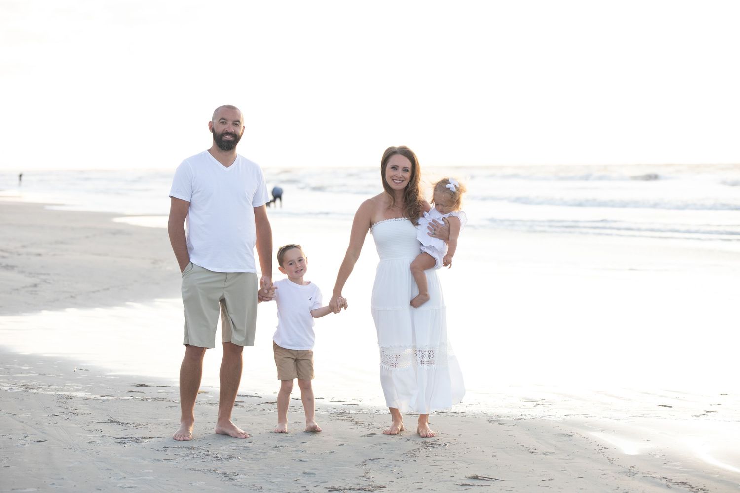 Family in white outfits walking together on sunny beach at sunset.