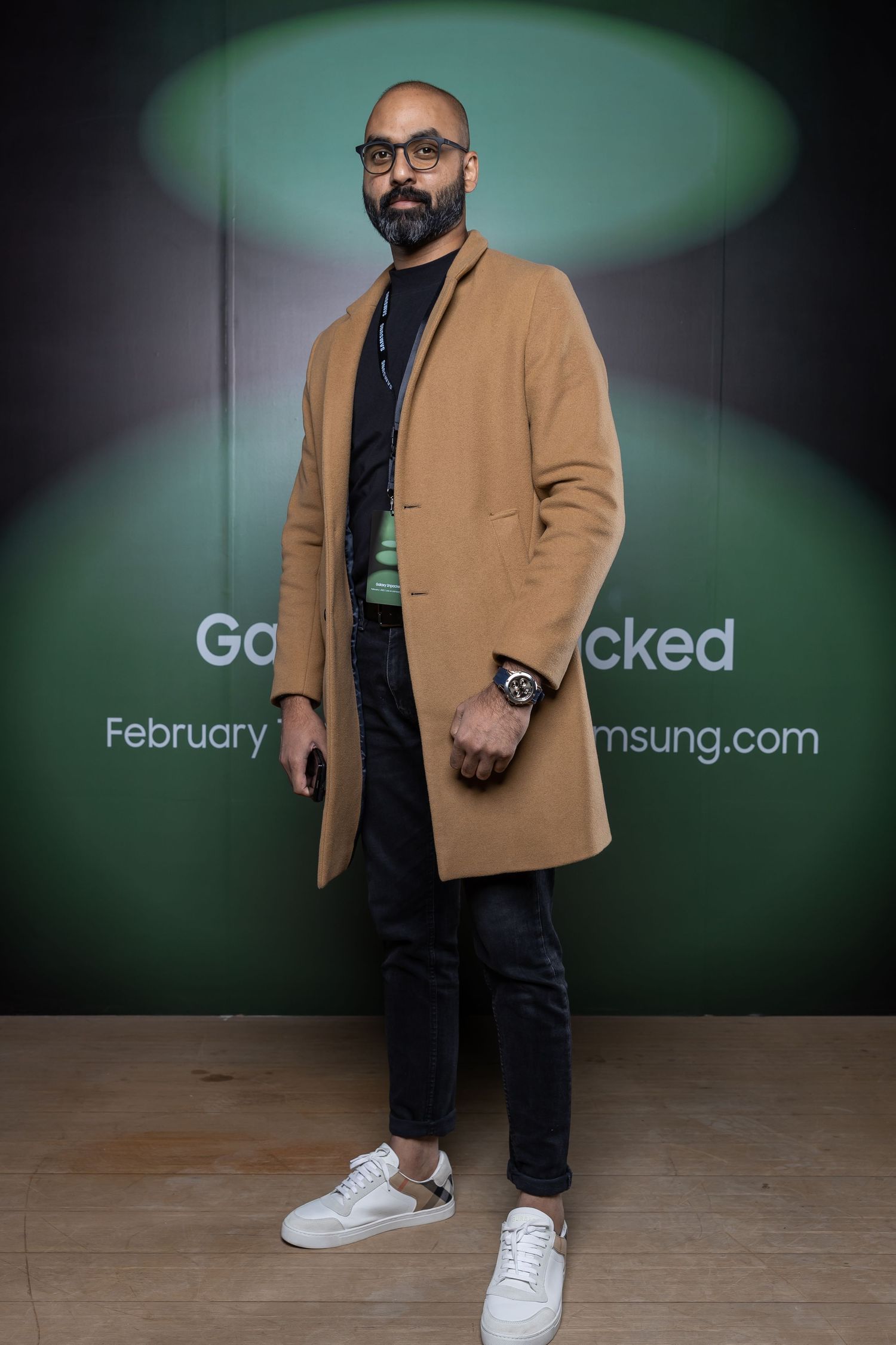 Stylish man wearing a camel coat with glasses standing against green backdrop at Samsung event.