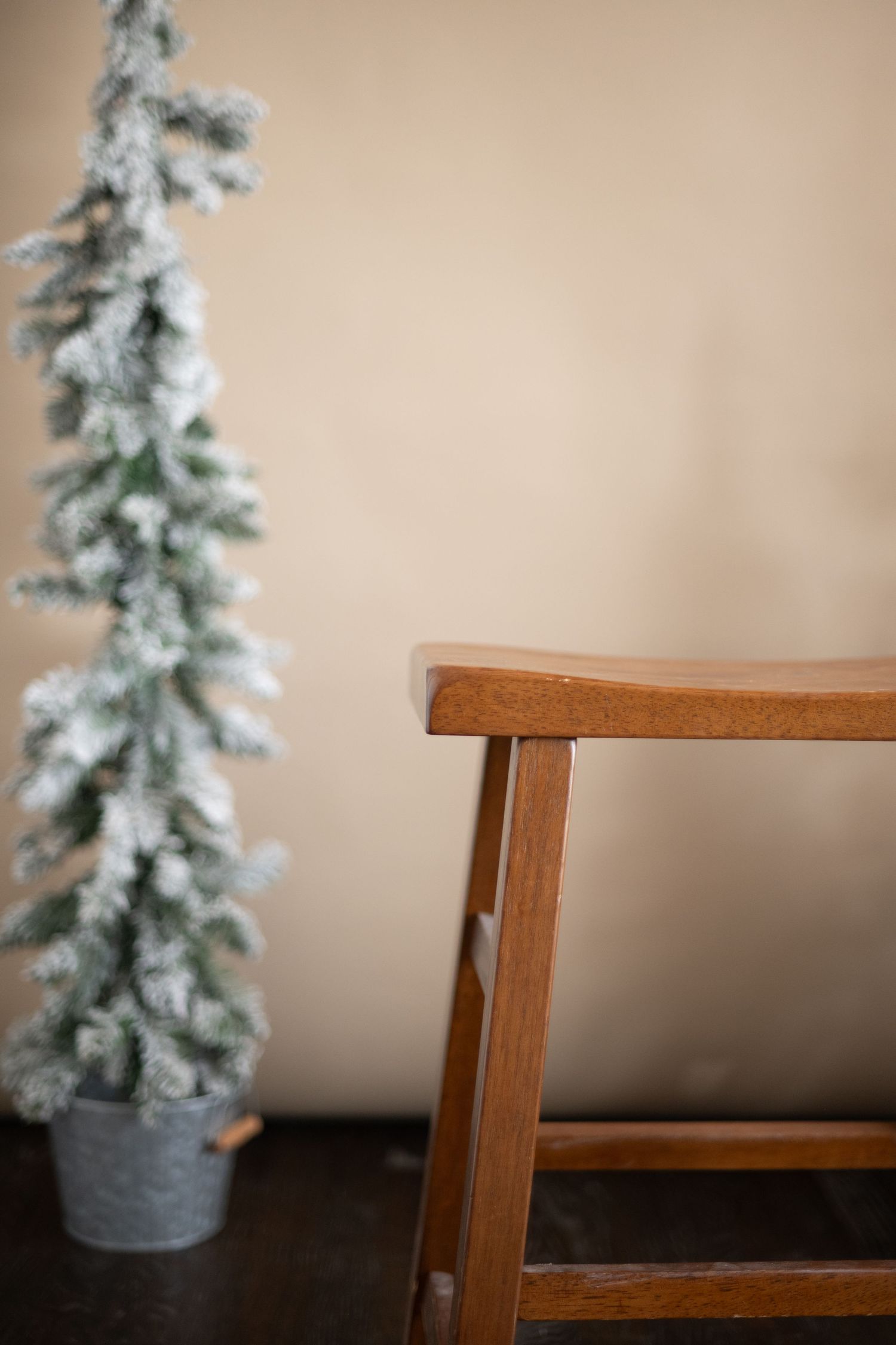 Minimalist holiday decor with flocked artificial pine tree next to wooden chair against neutral wall.