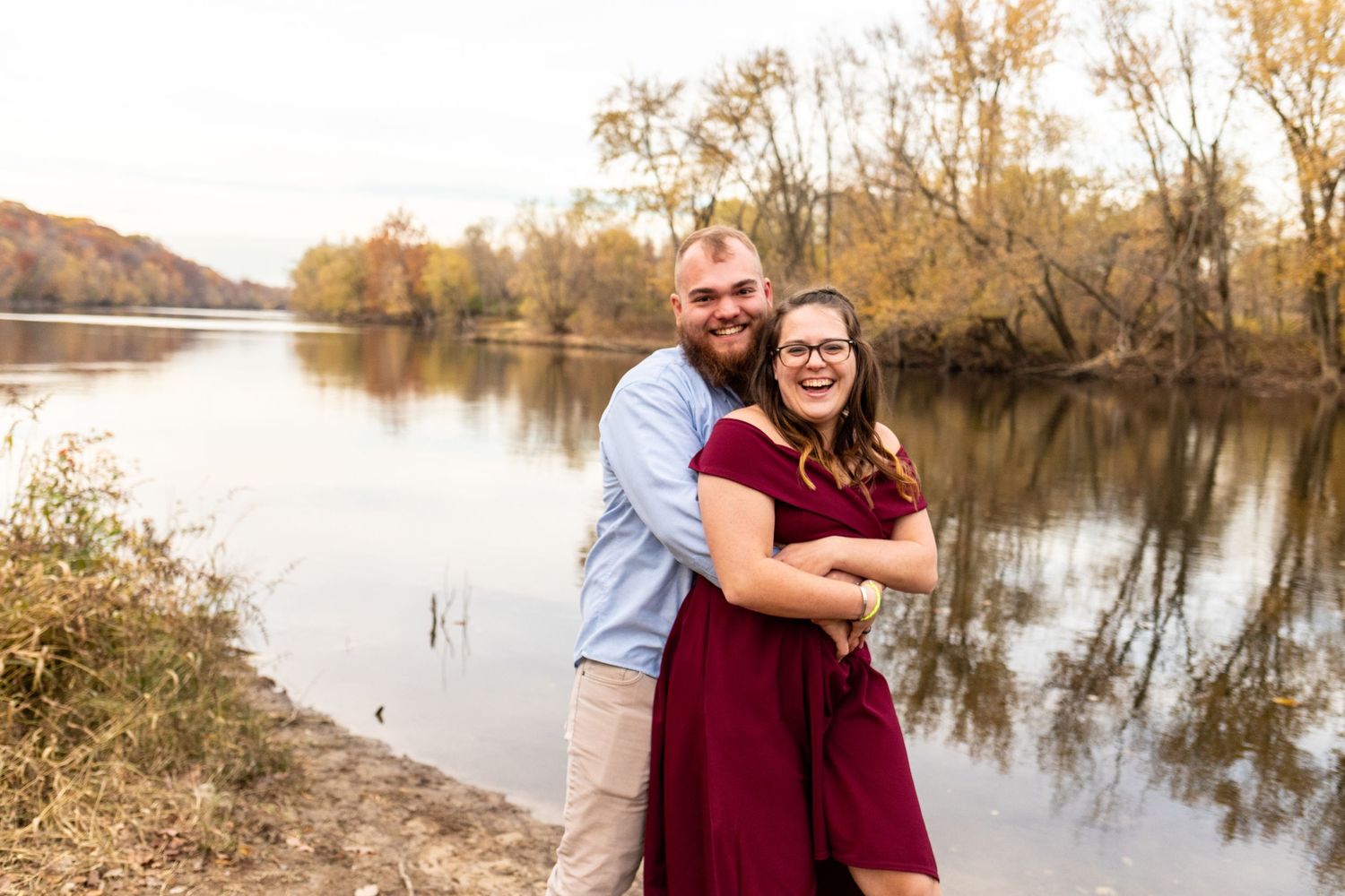 Happy couple embracing and smiling by a calm river surrounded by autumn foliage.