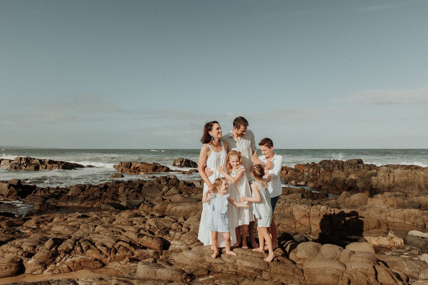 Family cuddle on a rocky platform by the beach with blue sky in the background