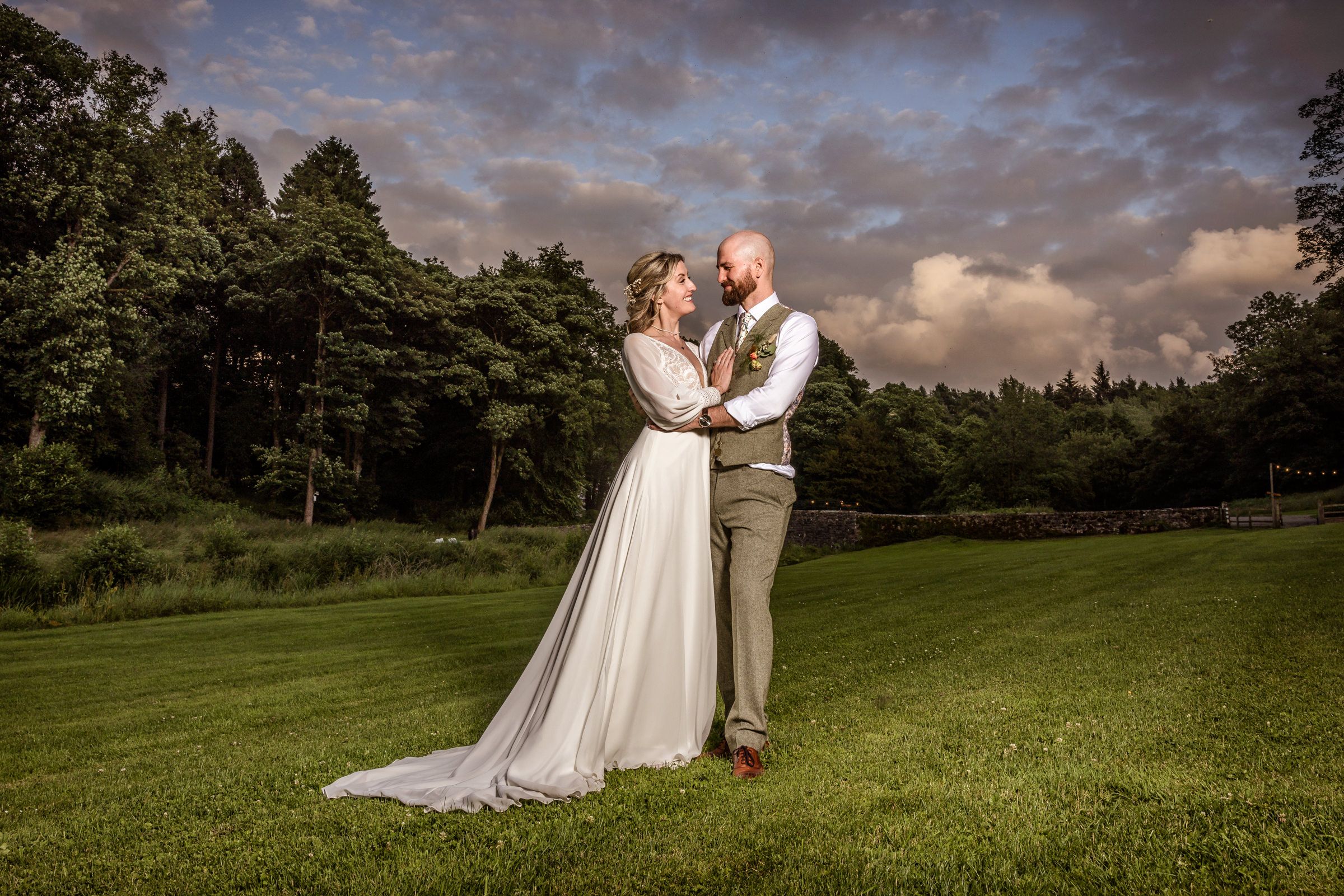 A romantic sunset wedding photo of a couple embracing on a lush green lawn with forest backdrop.