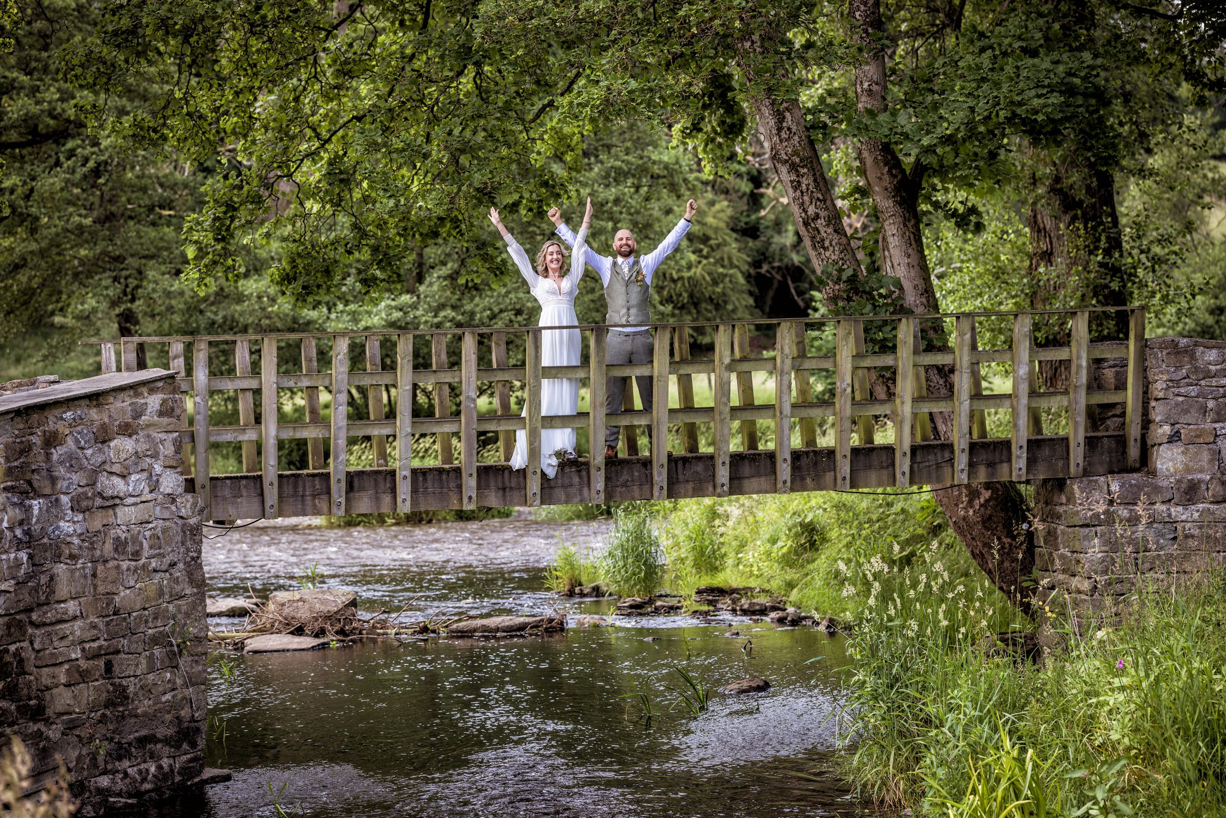 Joyful wedding celebration moment on a wooden bridge over a stream in a scenic woodland setting.