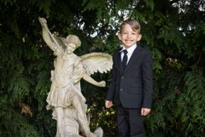 A child in a dark suit stands next to an angel statue in a garden setting.