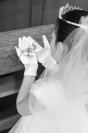 Close-up of white gloved hands holding a rosary with veil and tiara visible.