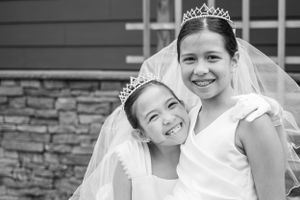 Two children in white dresses and tiaras share a special moment.