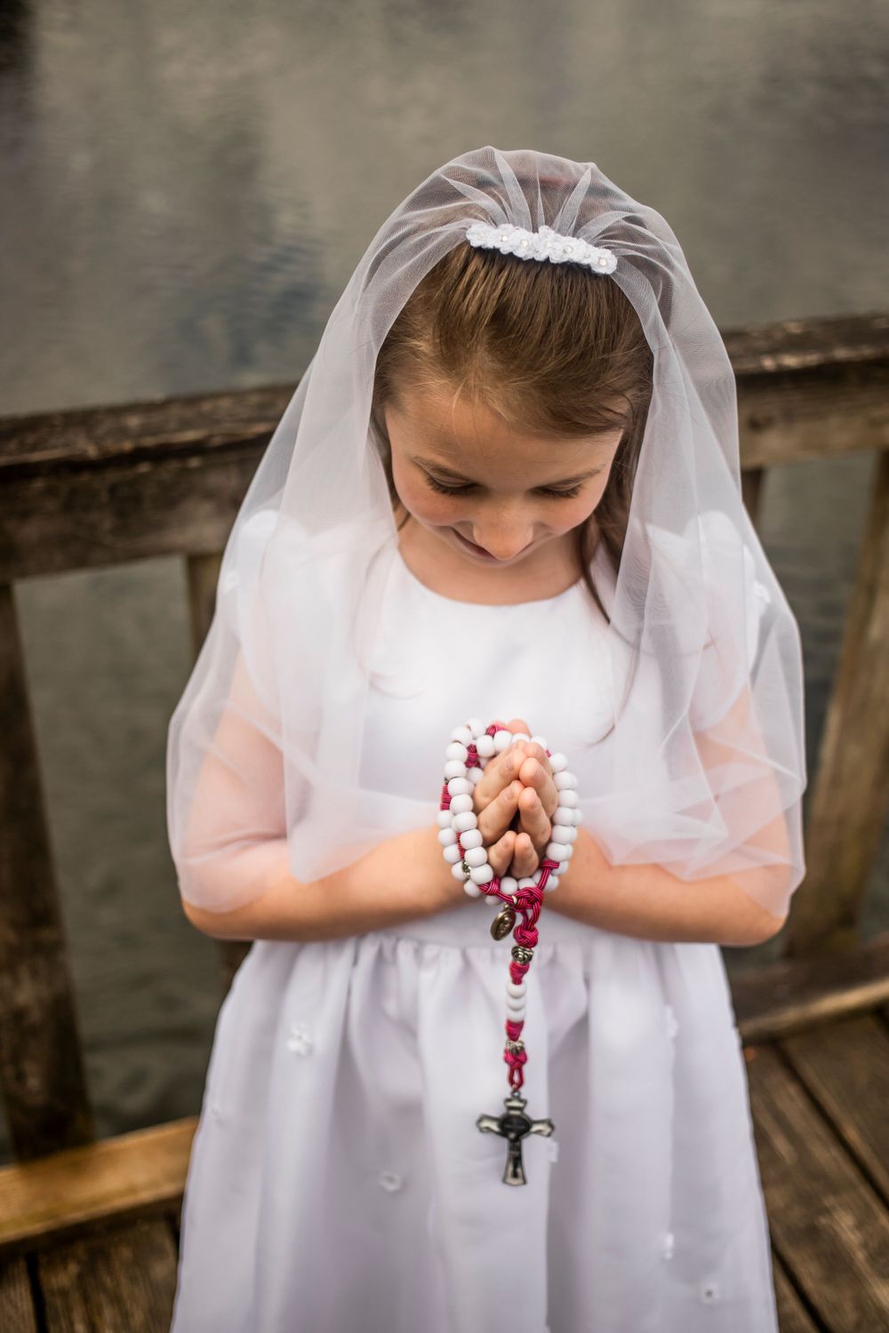 A figure in white holds rosary beads while wearing First Communion attire.