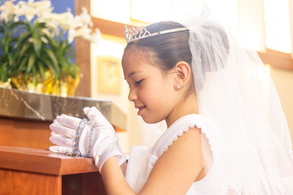 A child in white dress and veil prays in church.
