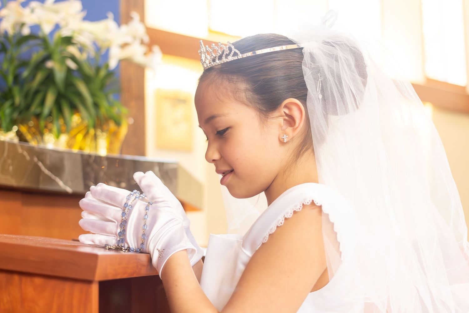 A child in white dress and veil prays in church.