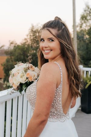 Bride in sparkling dress poses with bouquet on white deck railing at sunset.
