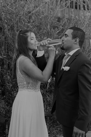 Newlyweds share a celebratory champagne toast in their wedding attire against a natural outdoor backdrop.