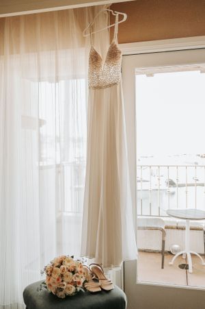 Elegant white wedding dress hanging by window with bouquet and shoes below.