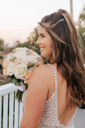 Bride in a sparkly white dress holds a peach and white floral bouquet while posing on a white porch railing.