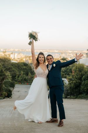 Joyful wedding couple poses with arms raised on a hilltop overlooking a city landscape at sunset.