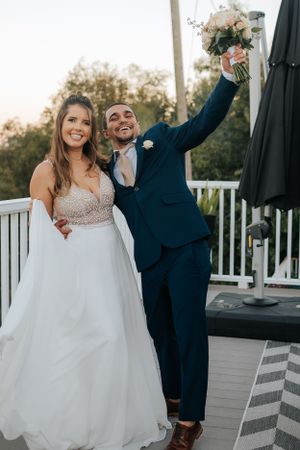 Newlyweds celebrate on outdoor deck as groom raises bouquet in celebration.