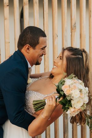 A joyful wedding couple laughing together while holding a bouquet of white and peach roses.