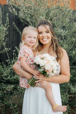 Bride in white dress holds bouquet while posing with young flower girl in floral dress.