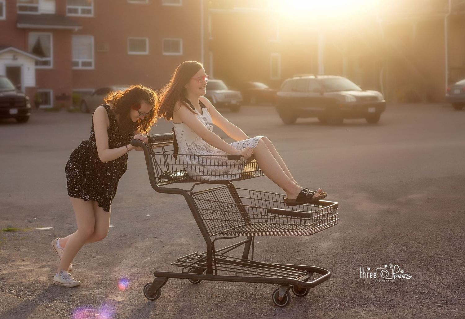 Girls playing with shopping cart in parking lot during golden hour sunset.