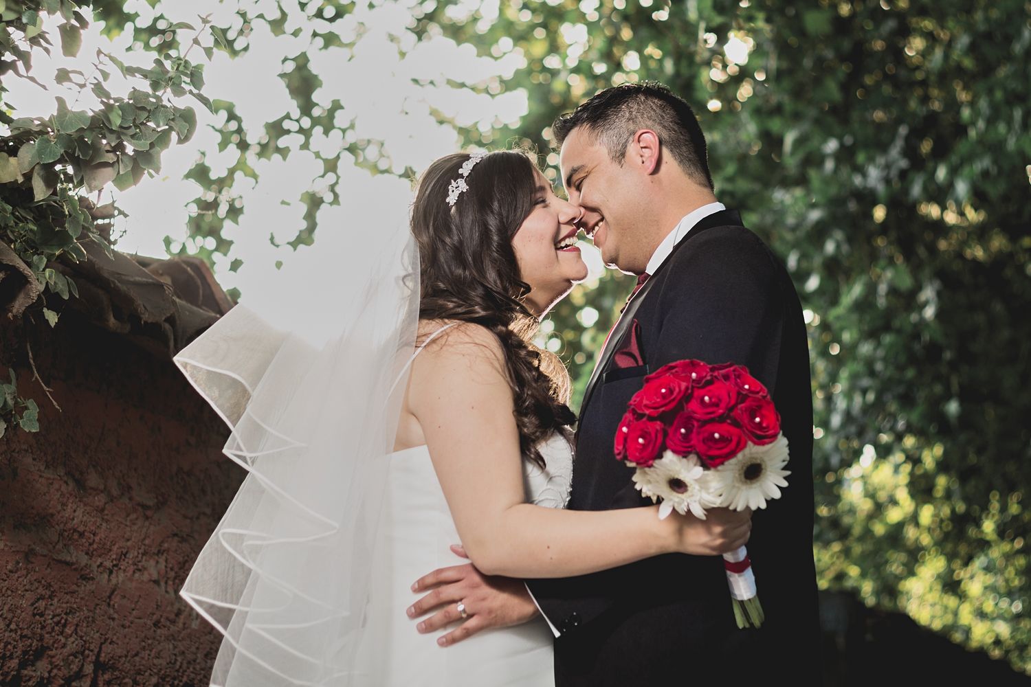 Intimate moment between newlyweds with red rose bouquet against natural backdrop.