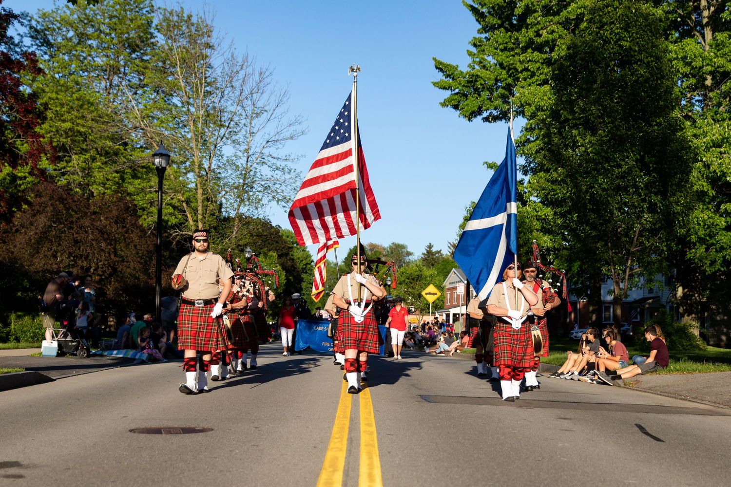 Parade participants in kilts marching with American and Scottish flags on sunny street.